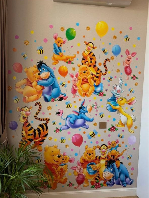 Adesivi murali di Winnie the Pooh e i suoi amici applicati su una parete beige di fianco a una pianta e un pavimento in legno.