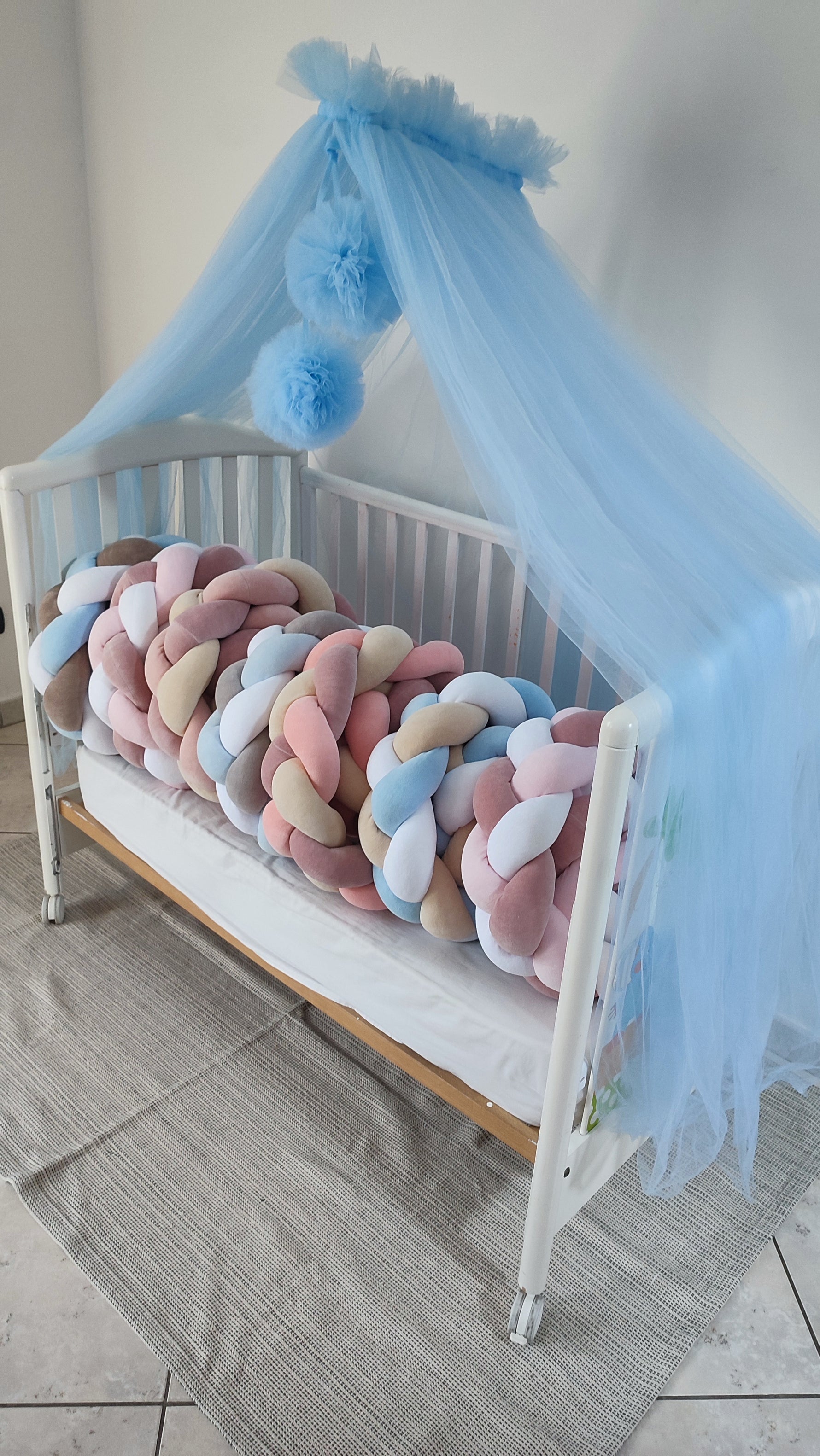 Varianti di paracolpi a treccia per lettino in diverse sfumature di rosa, azzurro e beige sotto un baldacchino in tulle.