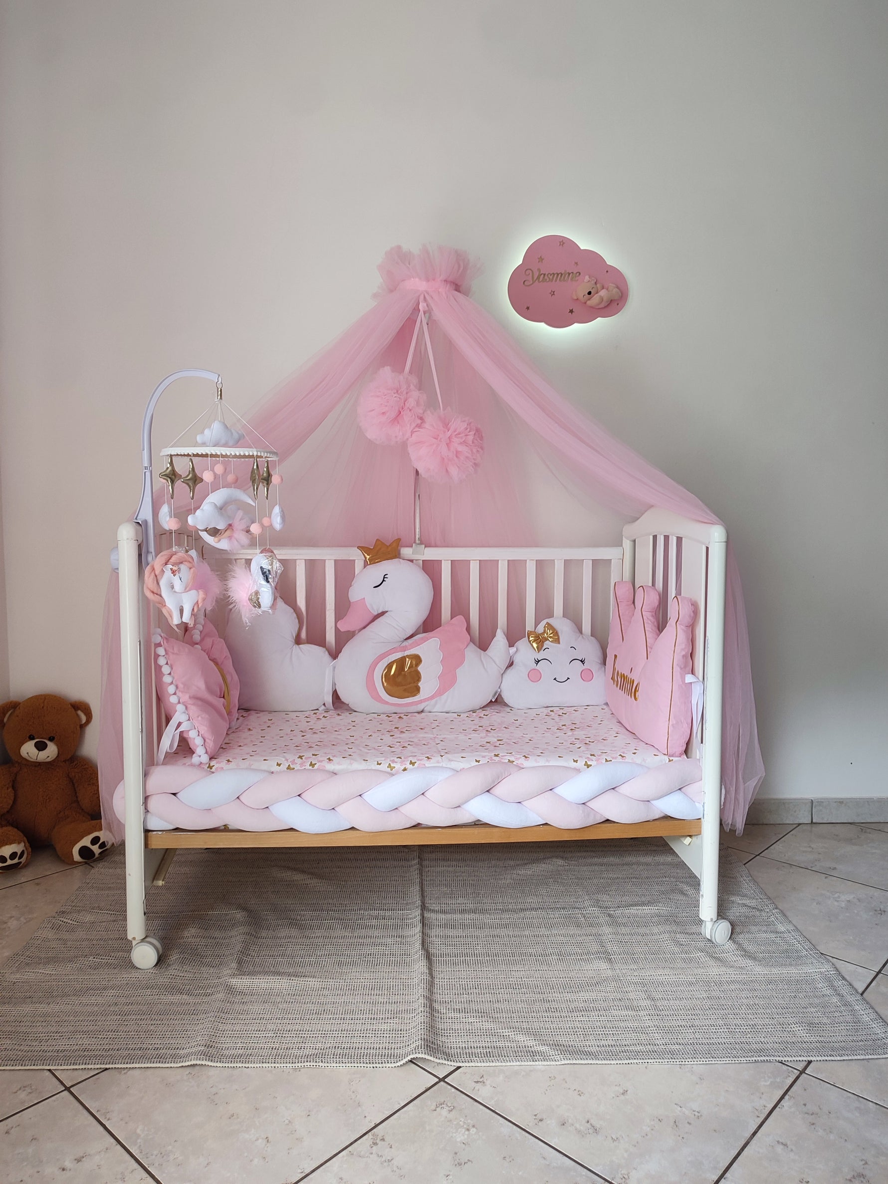 Lettino neonata rosa con baldacchino, paracolpi a treccia, giostrina mobile e cuscini decorativi a forma di cigno e nuvola.