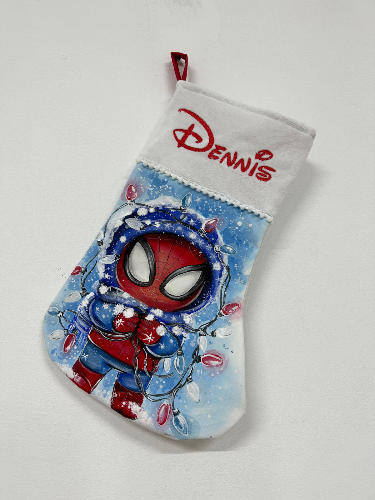 Calza di Natale azzurra dipinta a mano con illustrazione di Spiderman con cappuccio, avvolto da fili di luci natalizie.