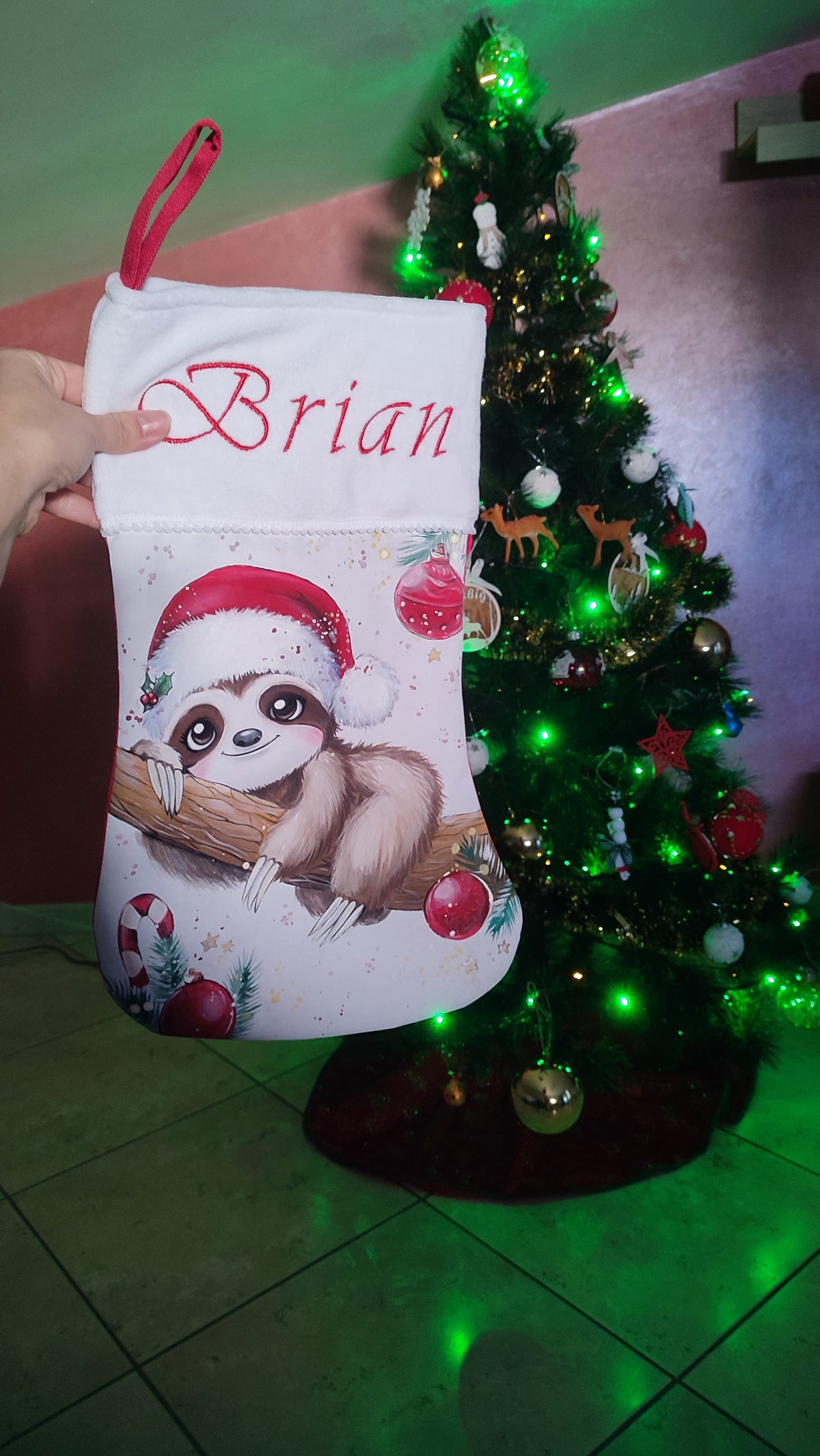 Calza di Natale bianca dipinta a mano con illustrazione di un bradipo con cappello di Babbo Natale e sfere rosse.