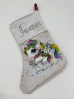 Calza di Natale grigia dipinta a mano con illustrazione di un unicorno dalla criniera arcobaleno su una slitta con regali.