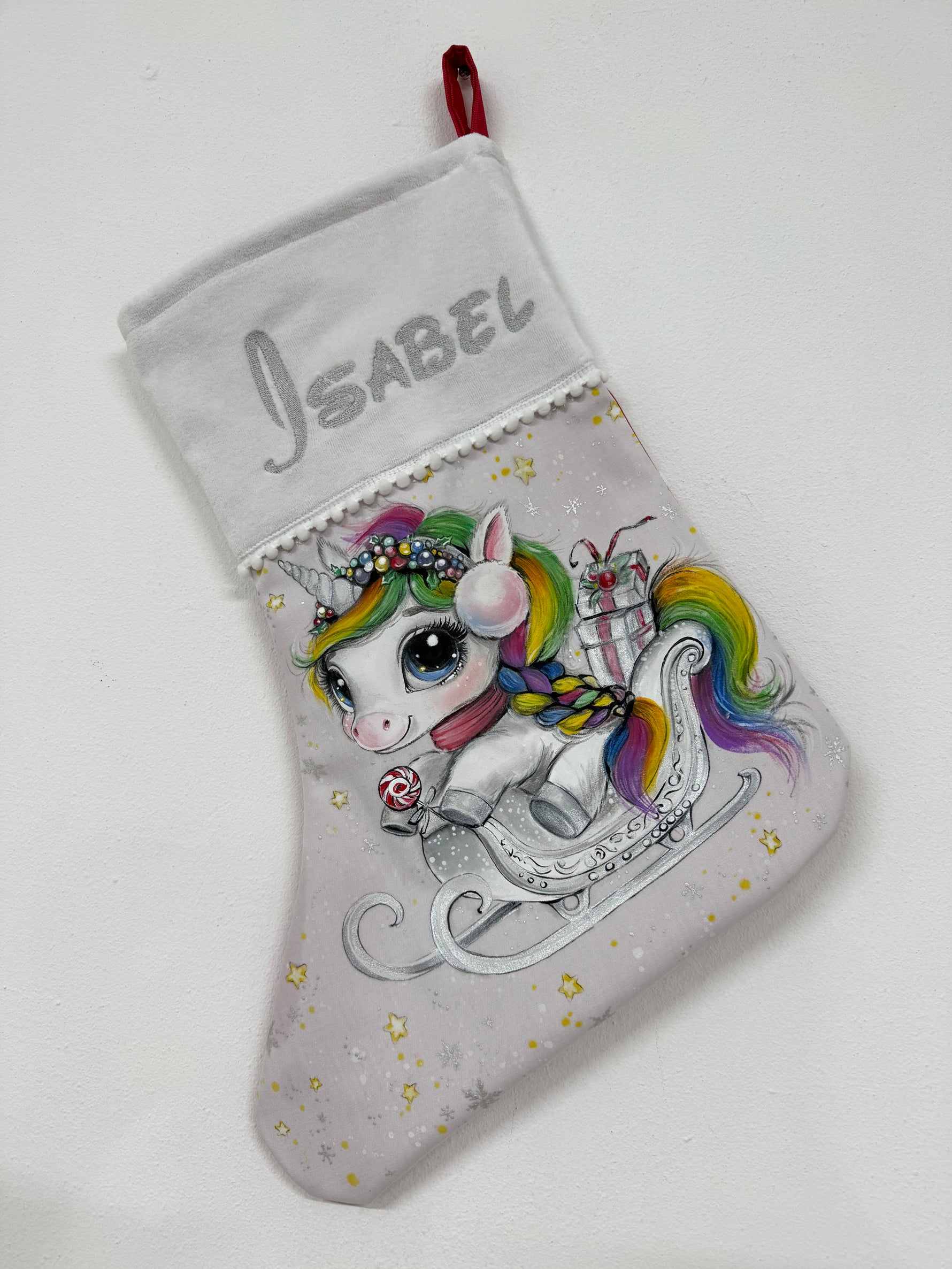 Calza di Natale grigia dipinta a mano con illustrazione di un unicorno dalla criniera arcobaleno su una slitta con regali.