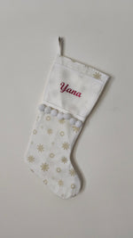 Calza di Natale bianca personalizzata con il nome Yana ricamato in rosso, stampe di fiocchi di neve oro e pom pom bianchi.