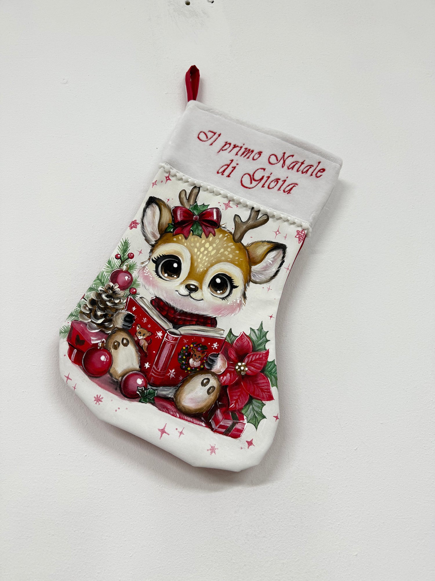 Calza di Natale bianca dipinta a mano per il primo Natale con illustrazione di una piccola renna, libro rosso e stella di Natale.