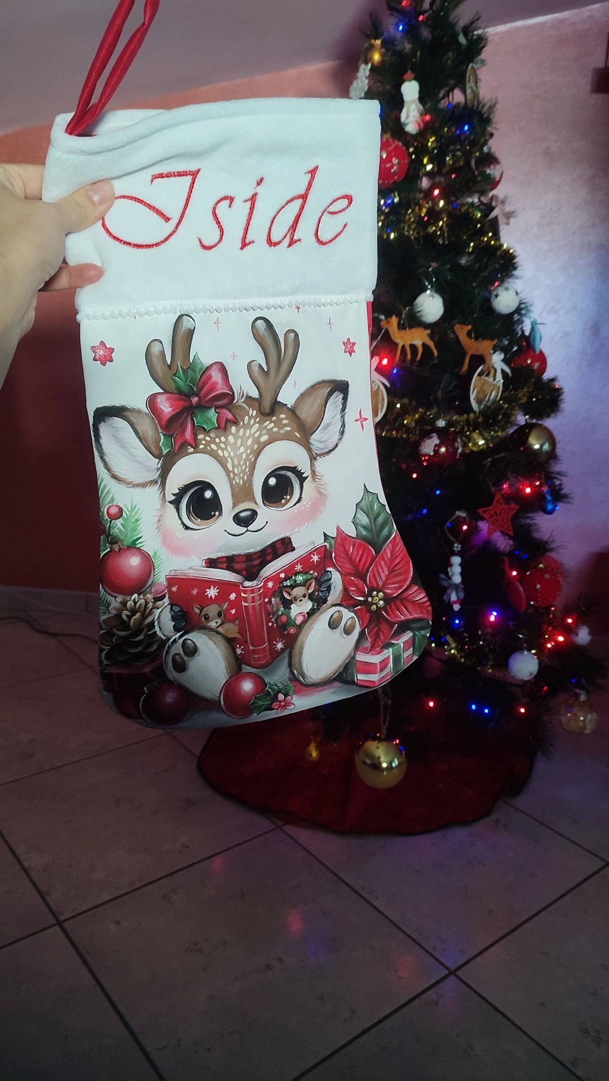 Calza di Natale bianca dipinta a mano con illustrazione di una piccola renna che legge un libro, pigne e una stella di Natale.