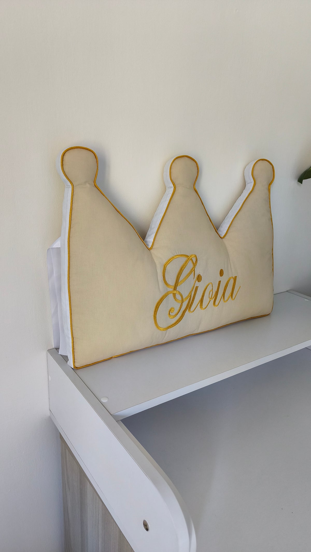 Cuscino paracolpi a forma di corona beige con ricamo personalizzato color oro, poggiato su una struttura letto bianca.
