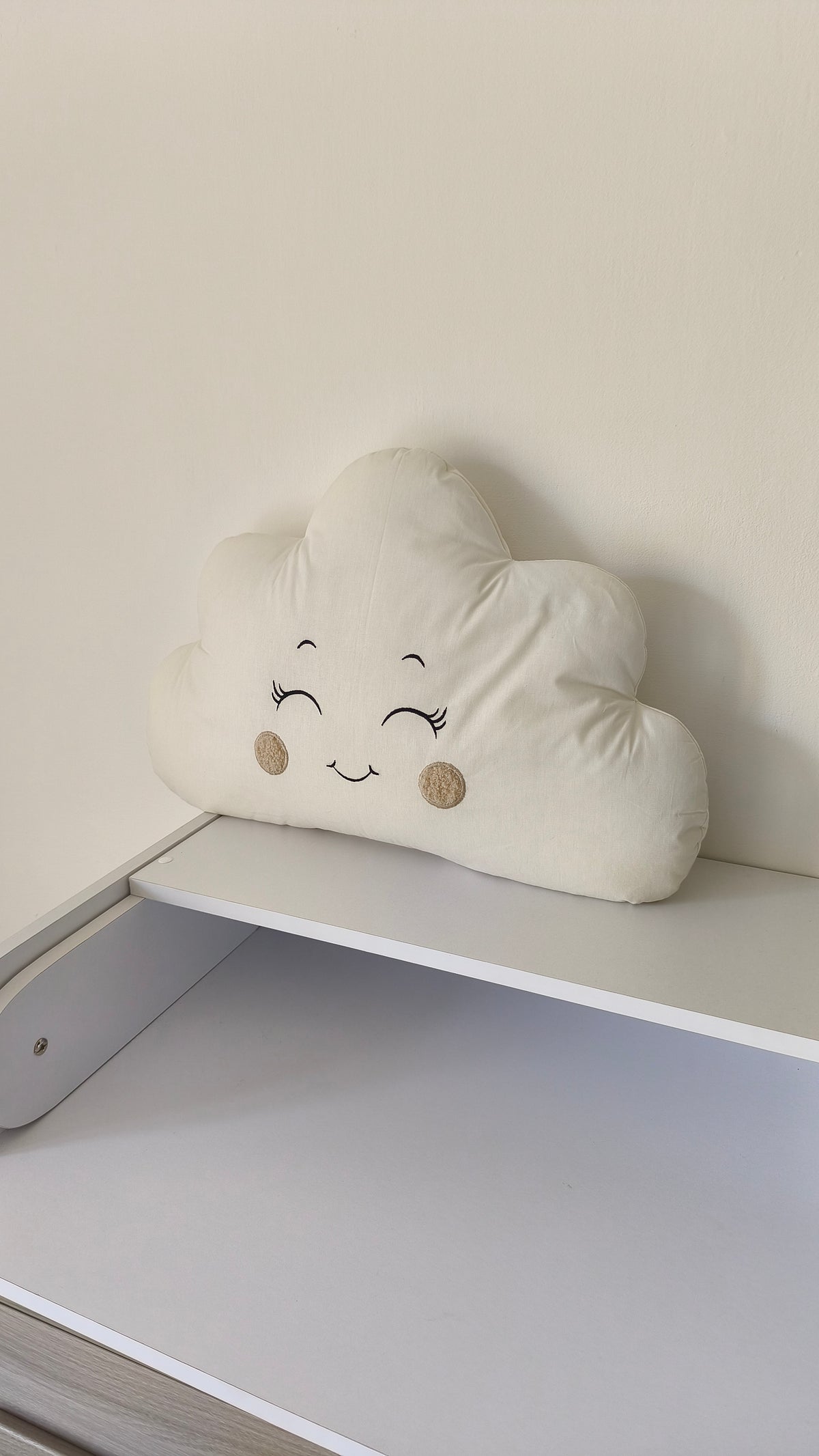 Cuscino paracolpi a forma di nuvoletta color panna con occhi ricamati e guance beige, ideale per una culla neutra.
