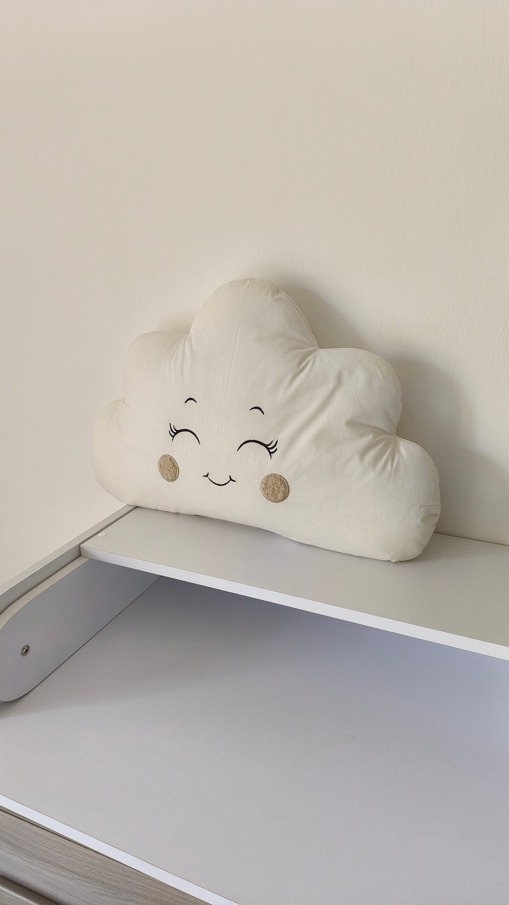 Cuscino paracolpi a forma di nuvoletta color panna con occhi ricamati e guance beige, ideale per una culla neutra.