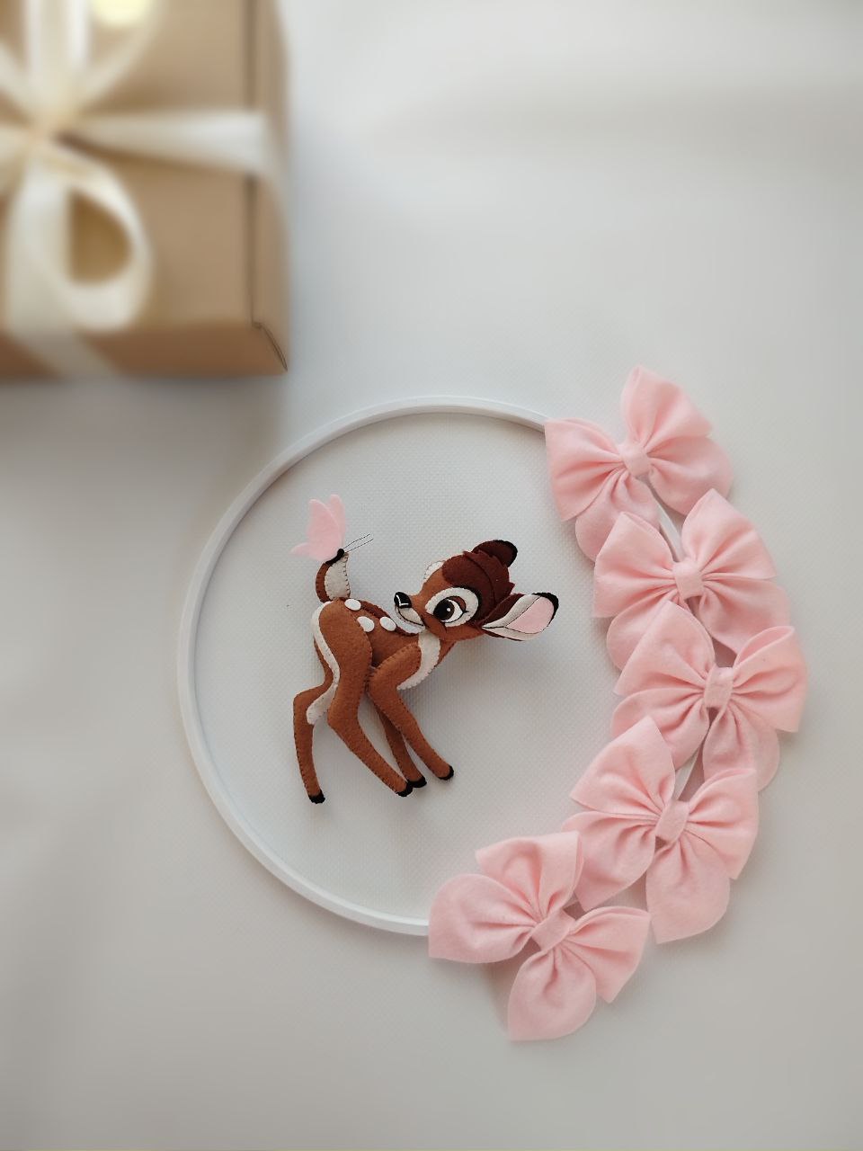 Dettaglio giostrina feltro con cerbiatto Bambi marrone, farfalla e grandi fiocchi rosa su sfondo bianco.