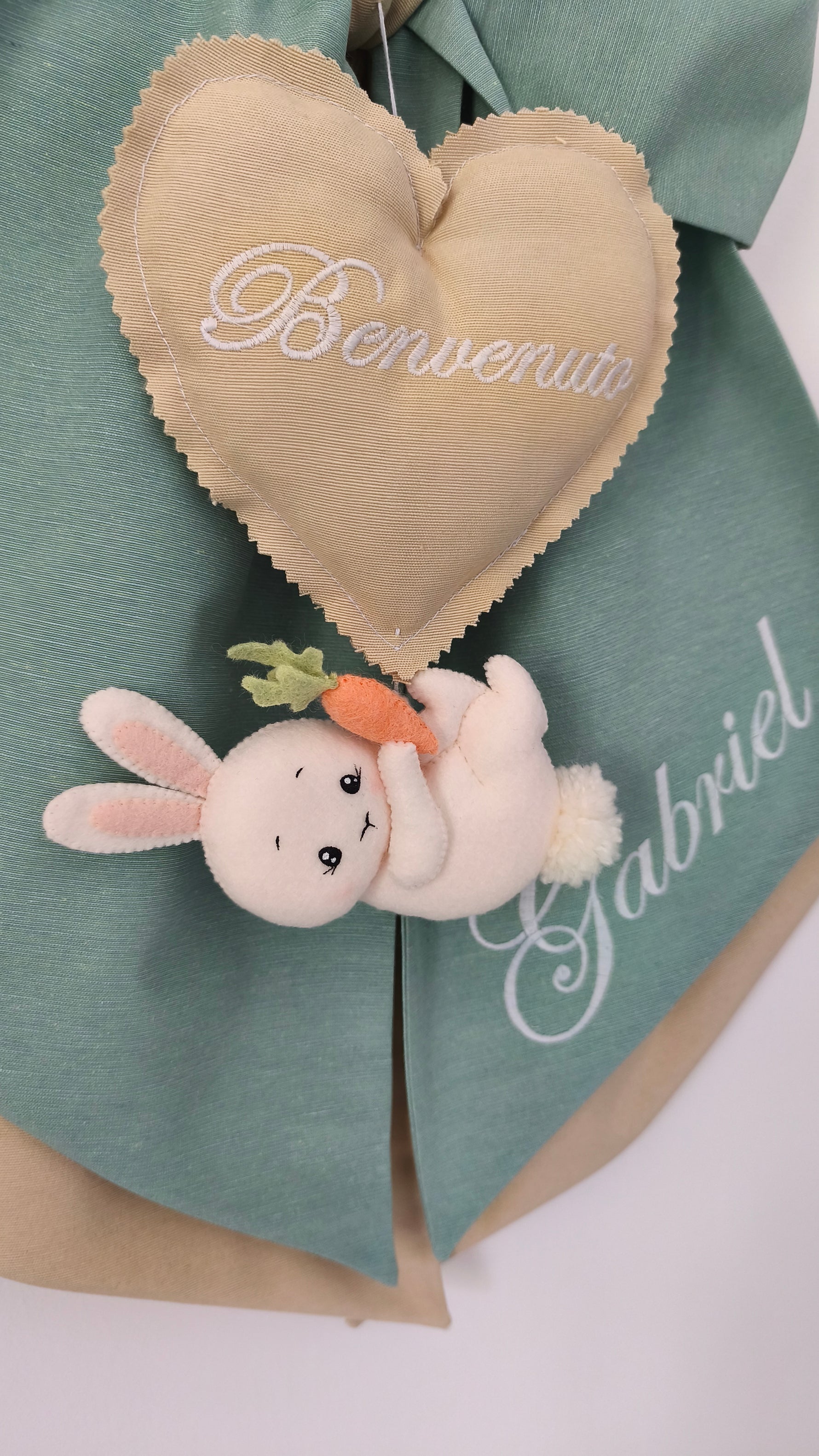 Dettaglio fiocco nascita con coniglietto bianco e carota in peluche, cuore beige ricamato benvenuto e nome sul tessuto verde salvia