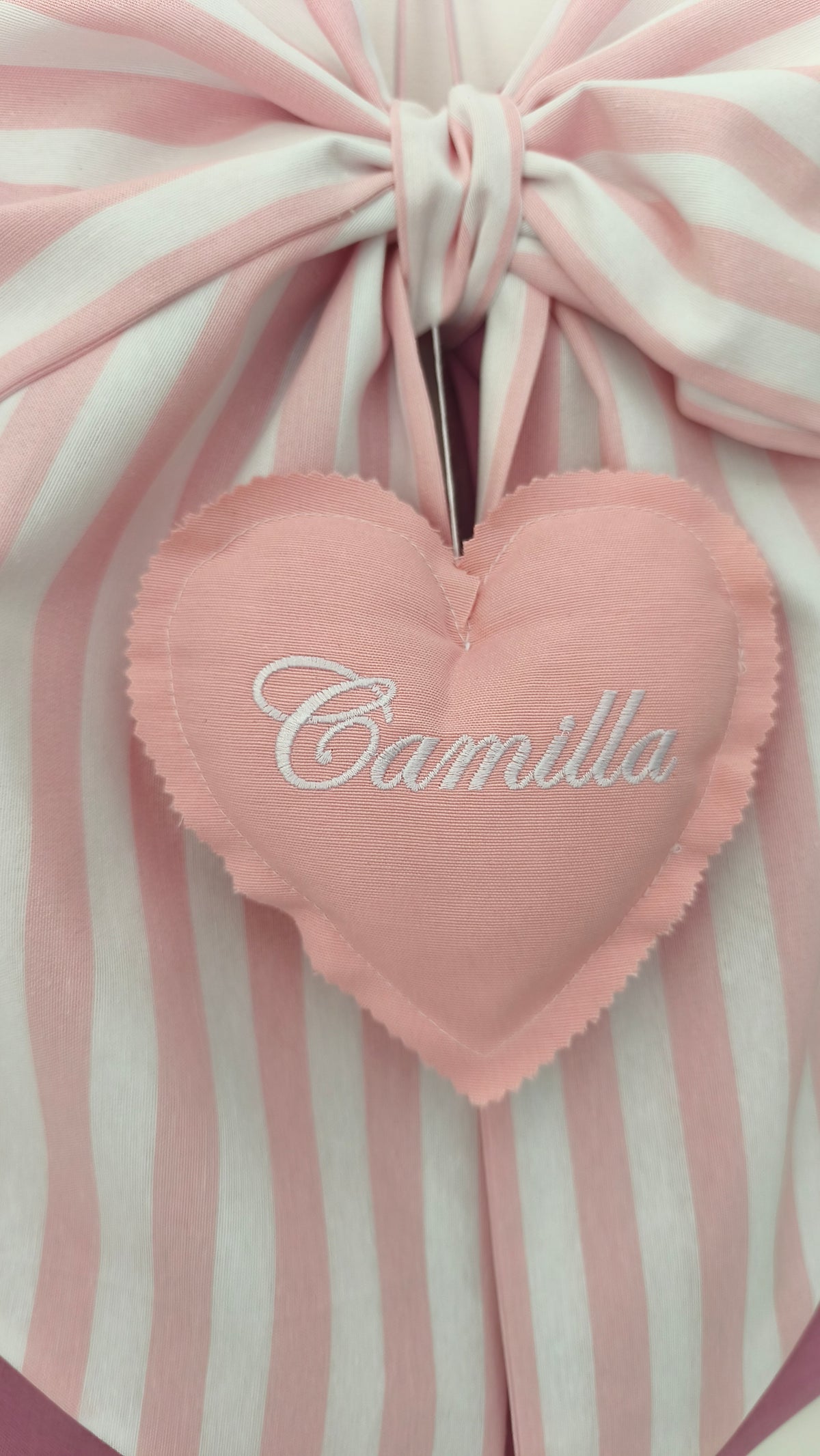 Dettaglio ravvicinato di un fiocco nascita rosa a righe con cuore pendente ricamato personalizzabile in tessuto.