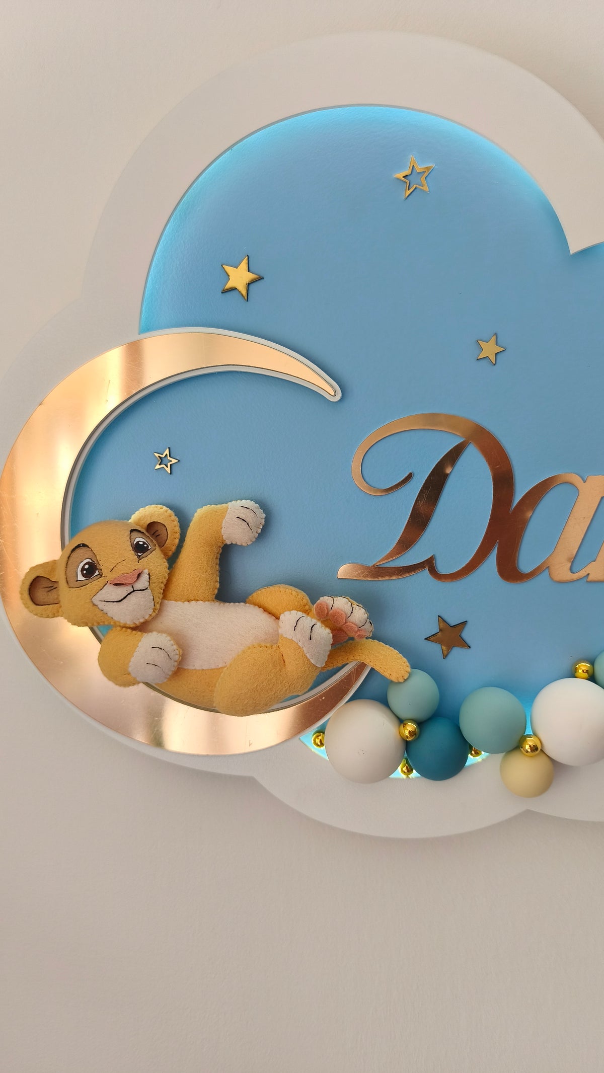 Dettaglio di lampada notturna neonato nuvola azzurra con Simba in feltro fatto a mano disteso sulla luna e palline decorative.