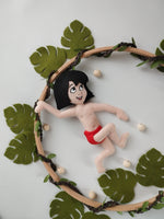 Dettaglio giostrina feltro con Mowgli in pantaloncini rossi su cerchio in legno decorato con foglie verdi. 