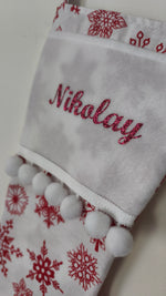 Primo piano del nome personalizzato ricamato con filo rosso glitterato su calza di Natale bianca morbida con pom pom bianchi.