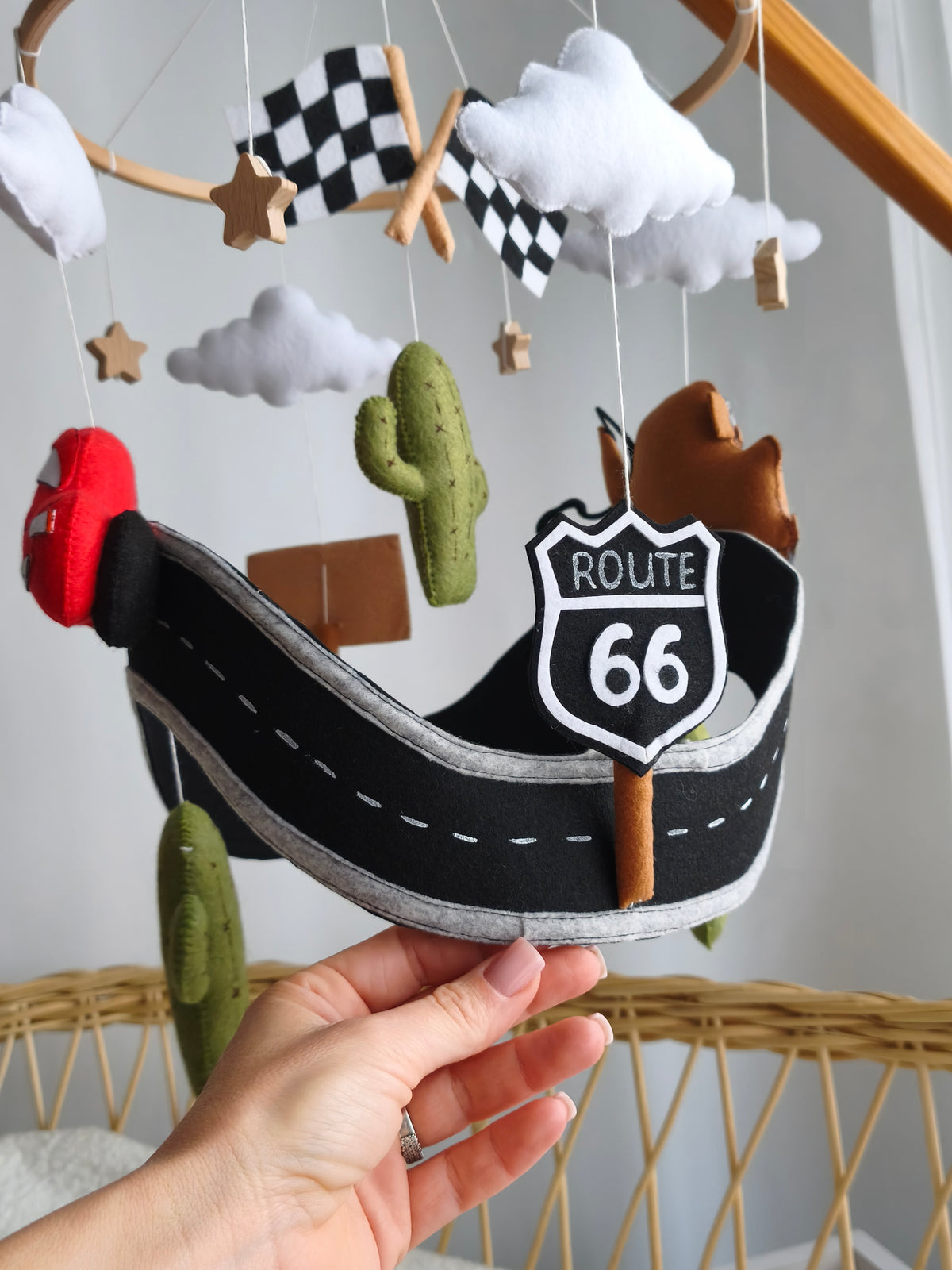 Dettaglio giostrina Cars feltro con cartello Route 66 nero, pista, cactus verde, bandiere a scacchi e nuvole.