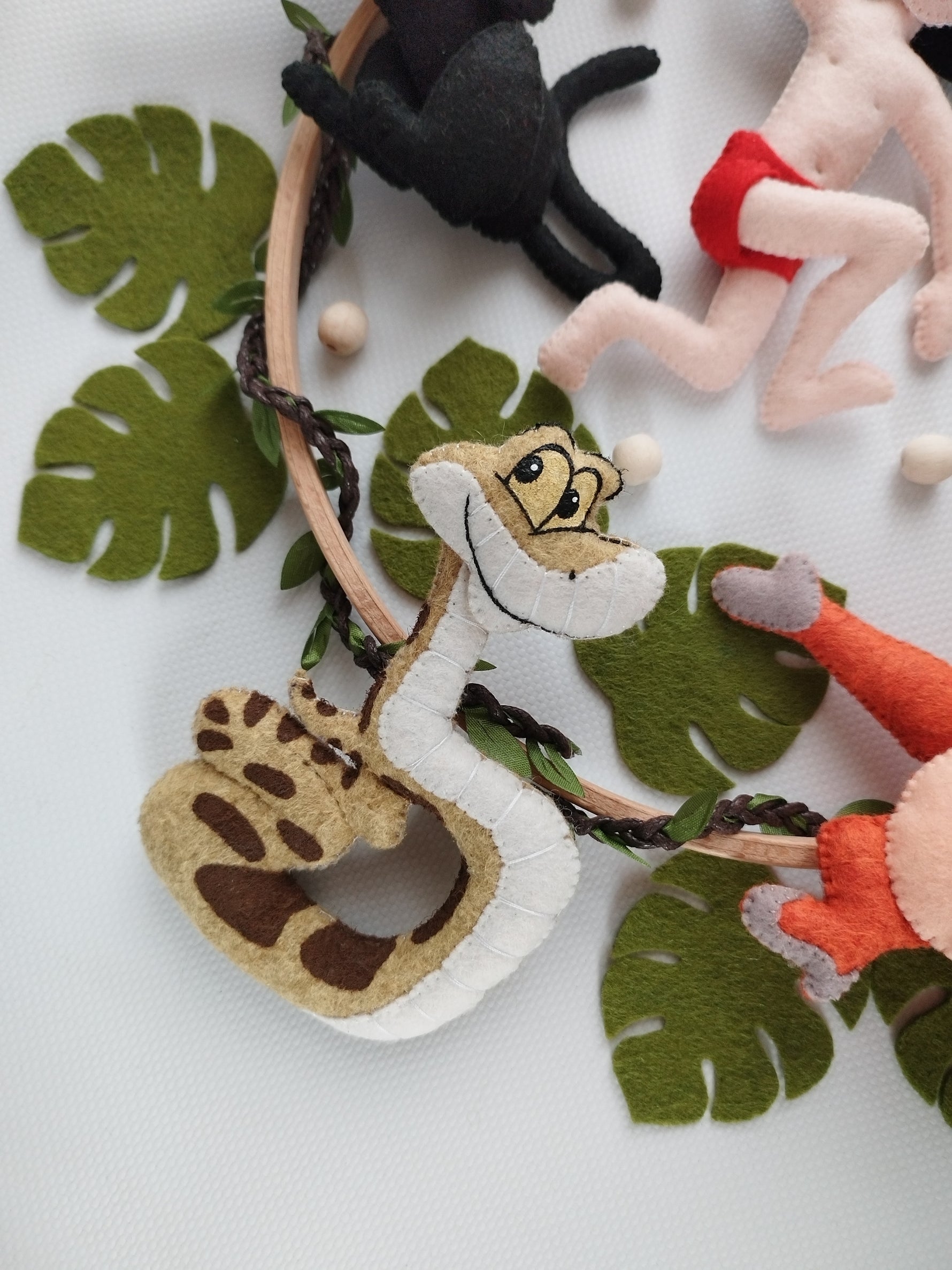Dettaglio giostrina musicale bambini feltro con serpente Kaa beige e marrone, foglie verdi tropicali e cerchio in legno. 