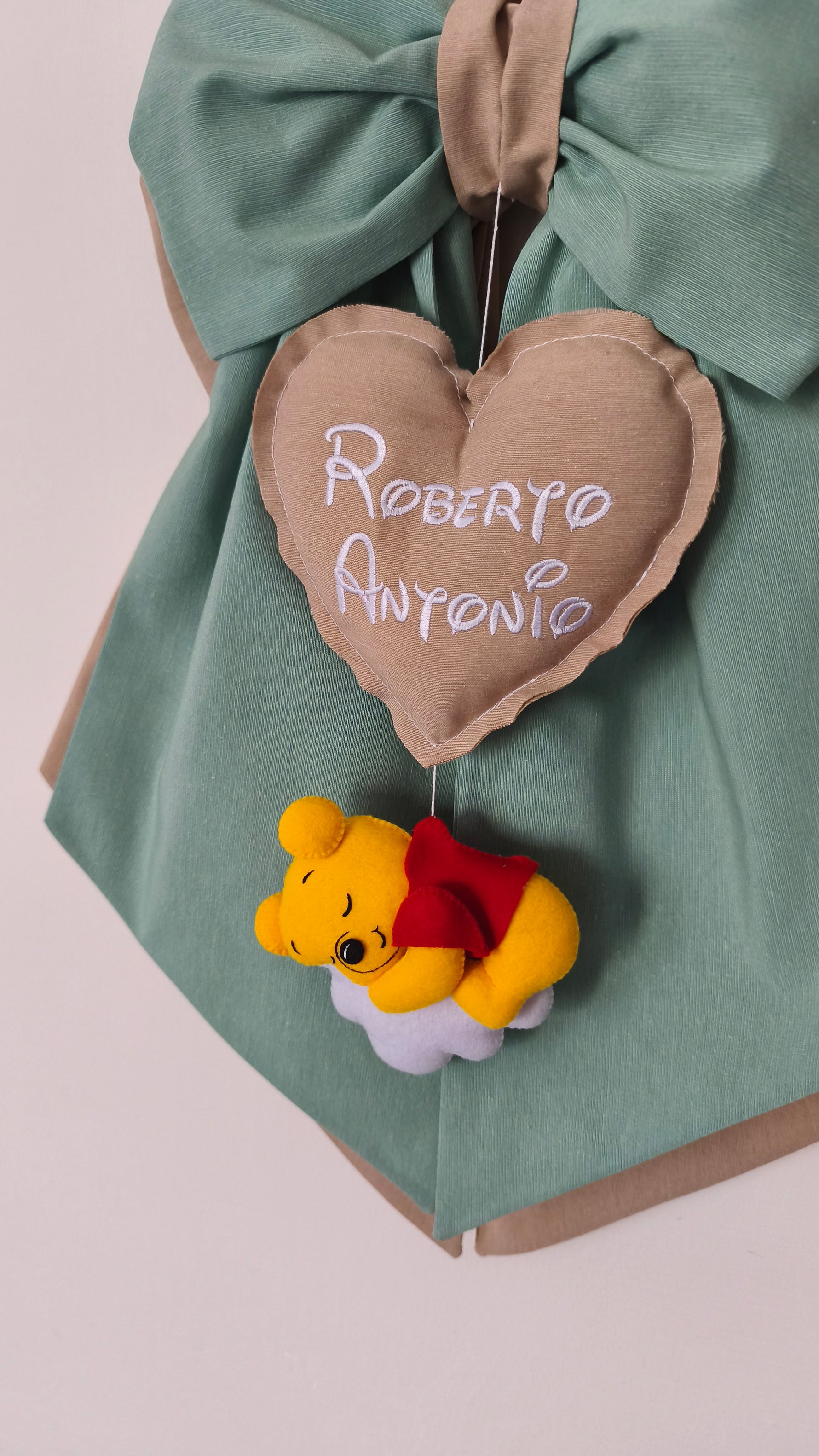 Primo piano di Winnie the Pooh in feltro su nuvola e cuore tortora con doppio nome ricamato per fiocco nascita bimbo.