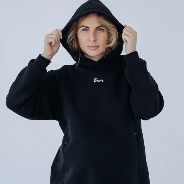 Donna sorridente indossa la felpa premaman nera Warm&Cosy tenendo il cappuccio, con ricamo bianco sul petto