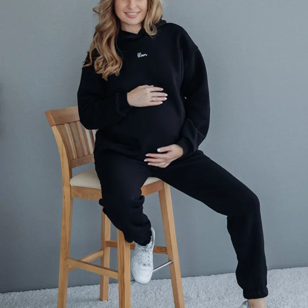 Donna in gravidanza seduta su uno sgabello con felpa premaman e allattamento nera con cappuccio