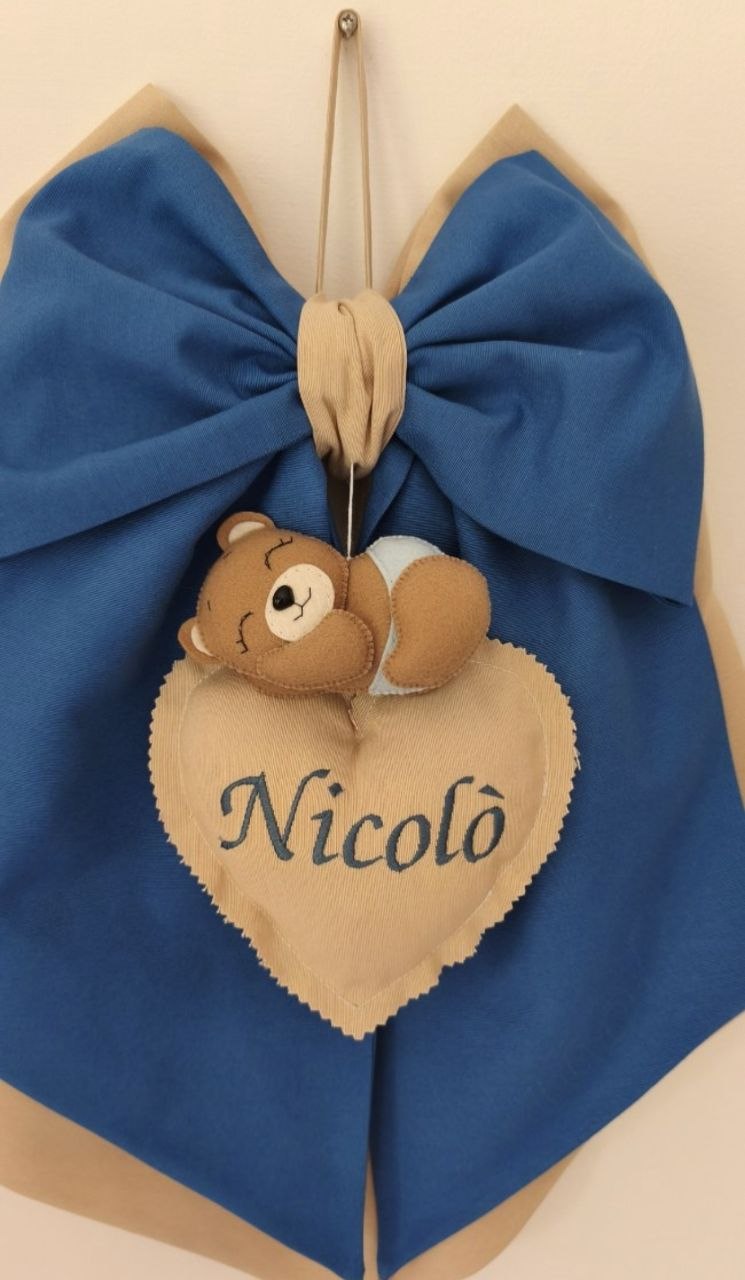 Dettaglio orsetto sopra il cuore in feltro con nome bambino, fiocco nascita blu da appendere alla porta