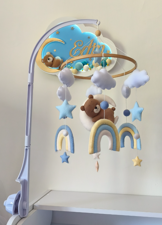Giostrina neonato in feltro con orsetto, arcobaleni azzurro e giallo e nuvole, appesa a un braccio in plastica bianca.