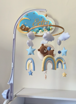 Giostrina neonato in feltro con orsetto, arcobaleni azzurro e giallo e nuvole, appesa a un braccio in plastica bianca.