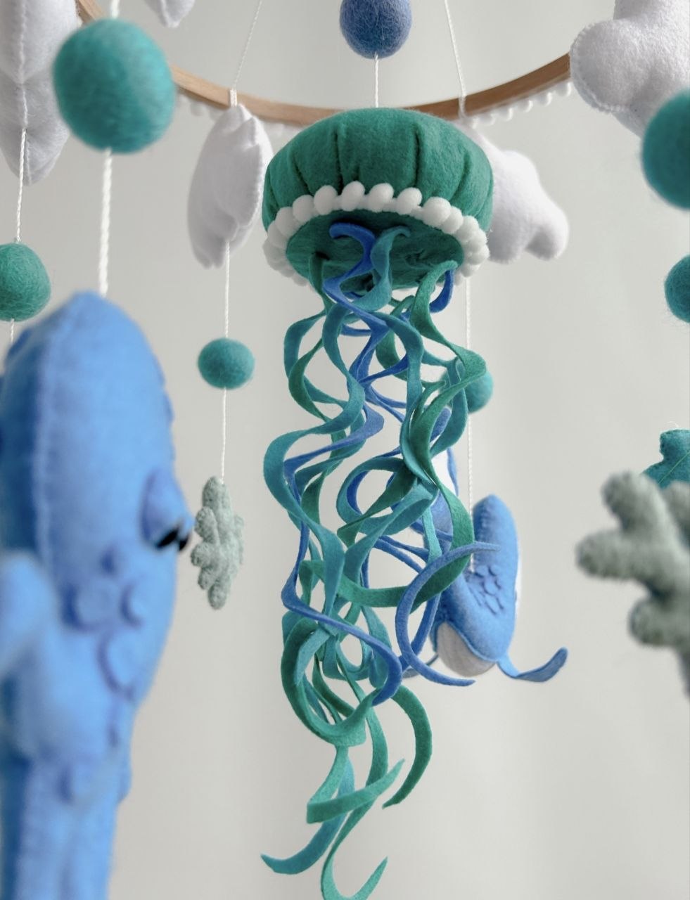 Primo piano medusa in feltro verde petrolio con tentacoli blu e turchese e pompon bianchi, giostrina culla mondo marino