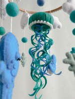 Primo piano medusa in feltro verde petrolio con tentacoli blu e turchese e pompon bianchi, giostrina culla mondo marino