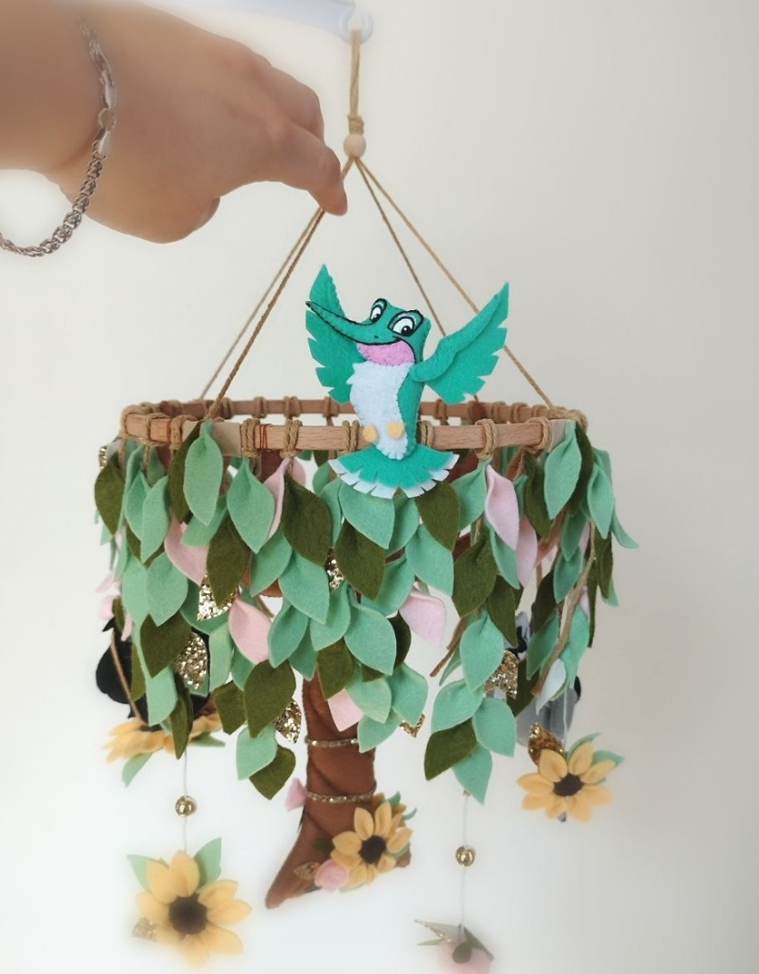 Giostrina culla Pocahontas in feltro con dettaglio del colibrì Flit in volo tra le foglie verdi e girasoli appesi.