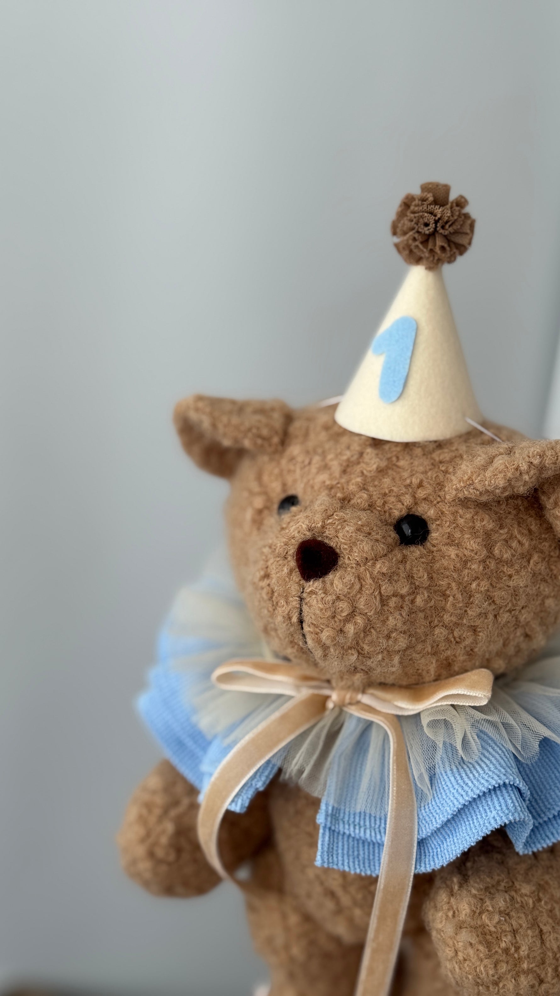 Orsetto di peluche beige con colletto a balze azzurro e cappellino col numero 1 per neonati