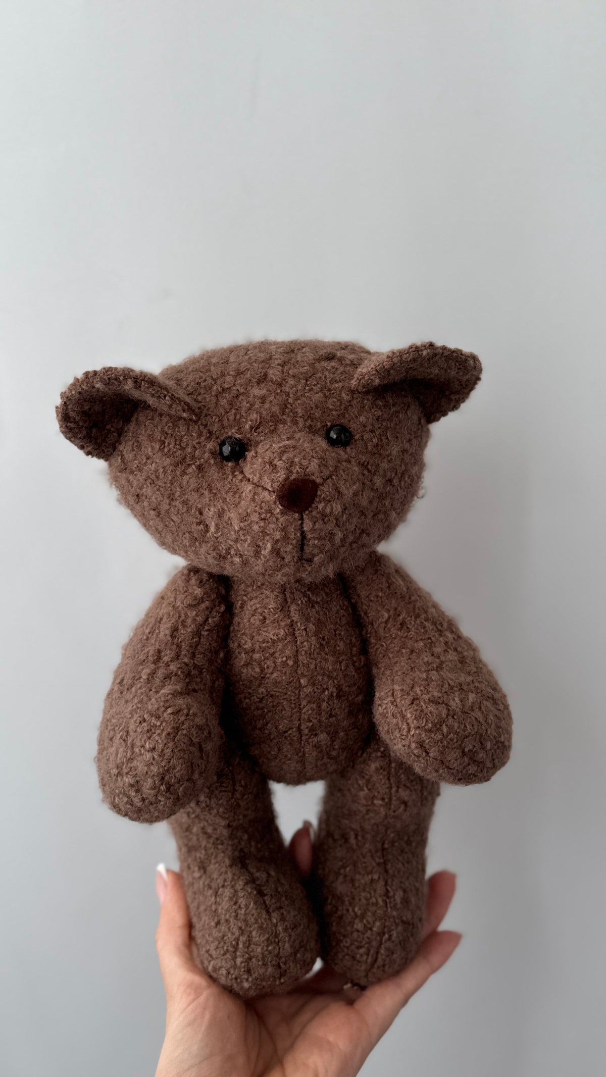 Orsetto di peluche marrone scuro in tessuto riccio bouclé per neonati sorretto da una mano