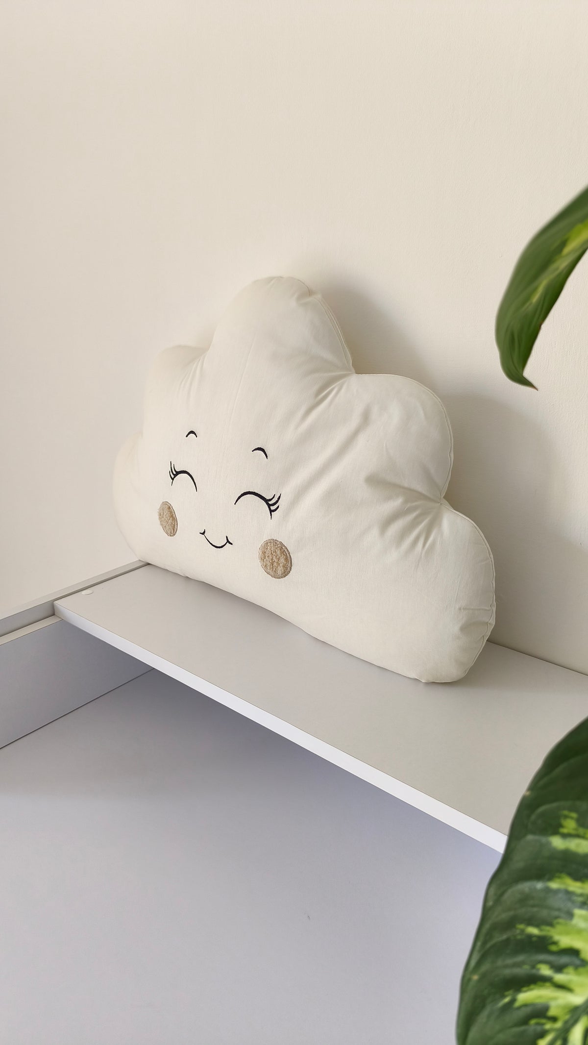 Primo piano di un cuscino paracolpi a forma di nuvola panna con occhi ricamati e guance beige per lettino neonato.