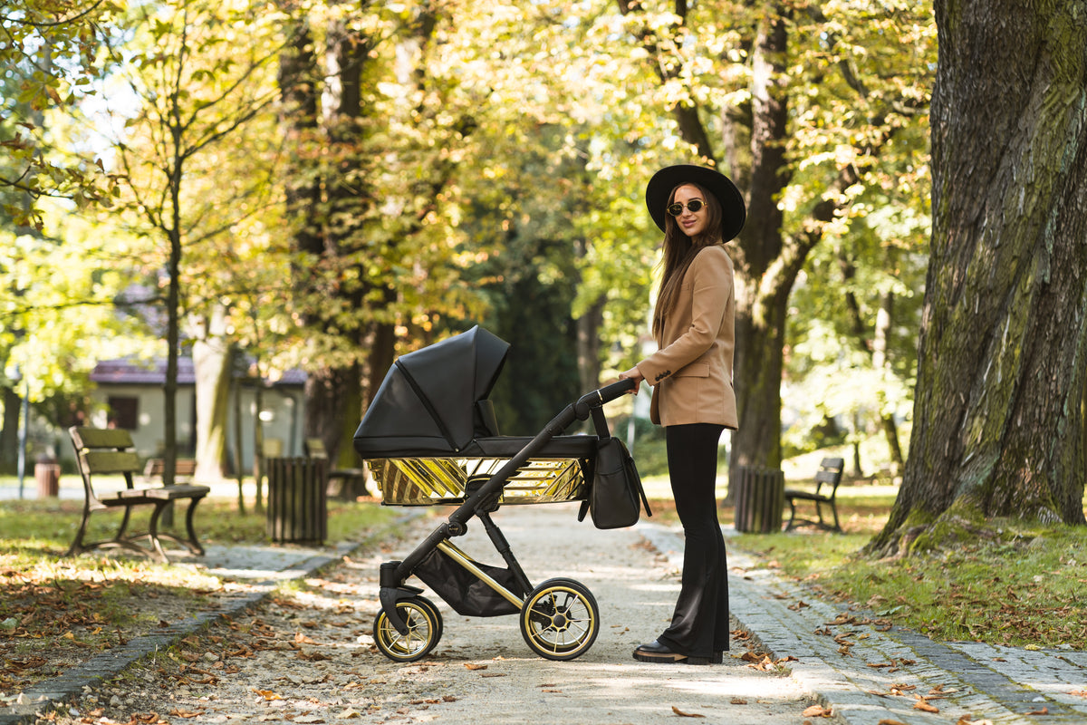 Mamma elegante con cappello passeggia nel parco con la carrozzina Kunert Ivento nera e oro lucido