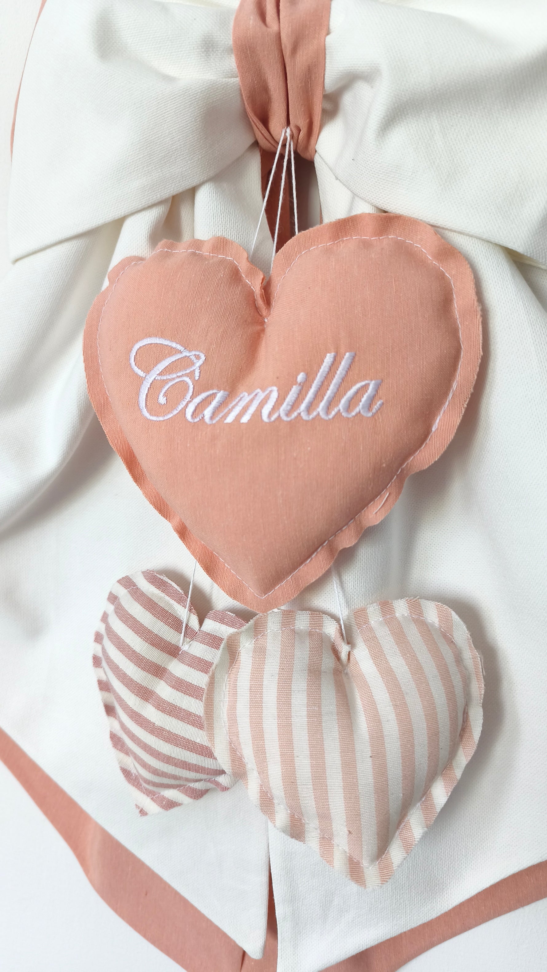 Primo piano coccarda nascita panna con cuori pendenti a righe e ricamo personalizzato su tessuto artigianale.