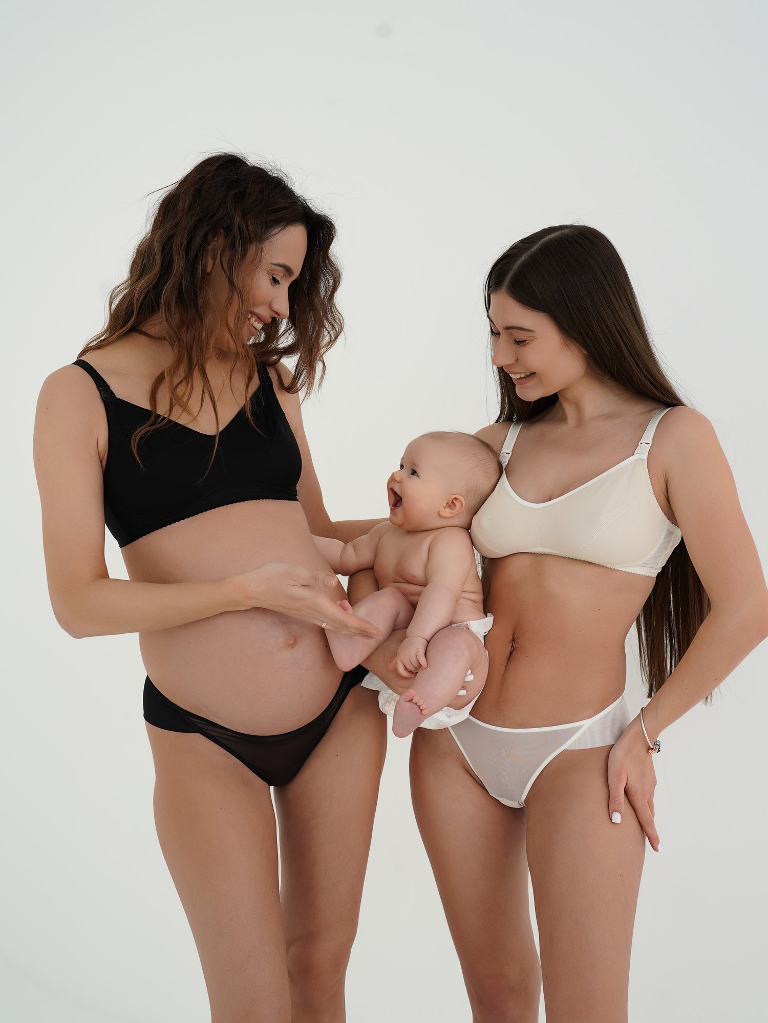Due donne in intimo premaman e allattamento, nero e beige, sorridono insieme a un neonato