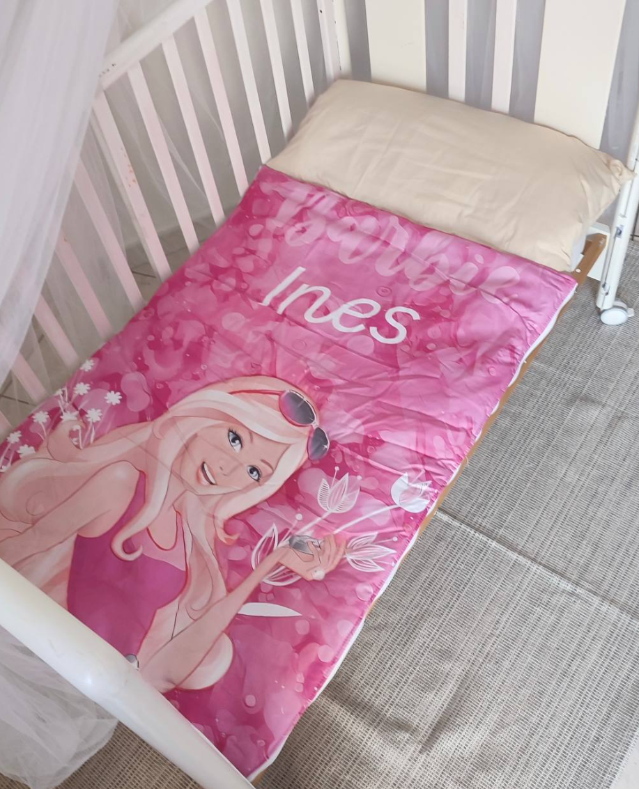 Sacco a pelo per bambini personalizzato con nome Ines, rosa con bambola bionda, steso in un lettino con cuscino beige