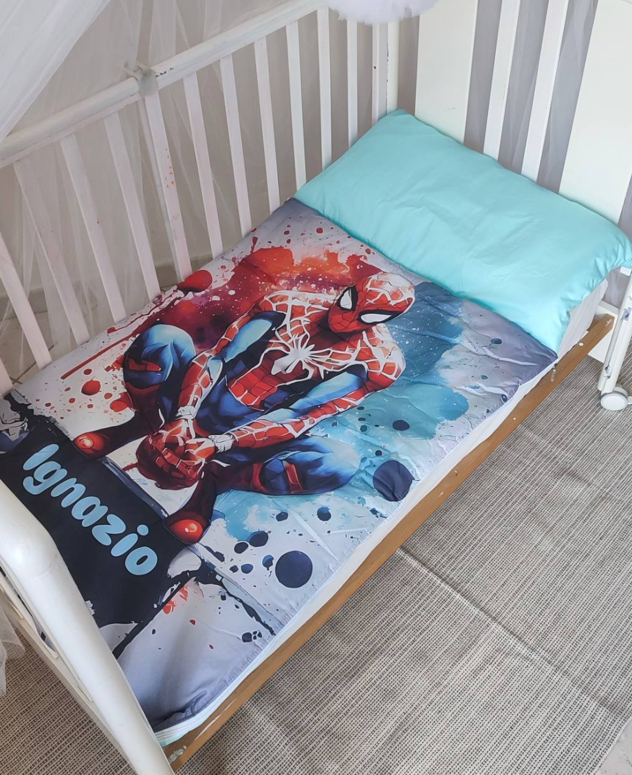 Sacco a pelo per bambini personalizzato con nome Ignazio, stampa supereroe rosso e blu, con cuscino azzurro, steso in un lettino bianco