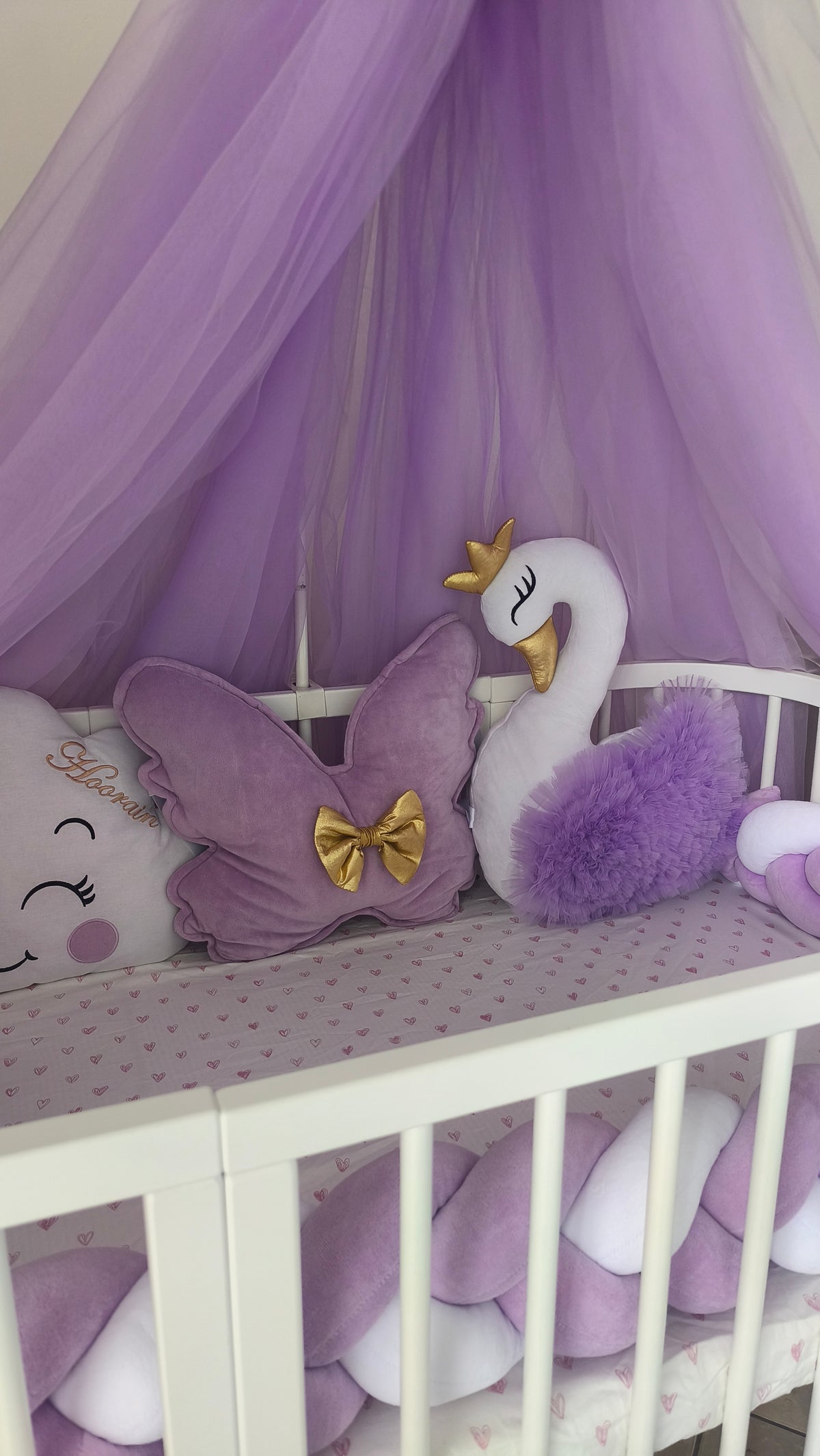 Dettaglio cuscino a nuvola con nome e fiocco viola, inserito in un set culla completo lilla e bianco con cigno.