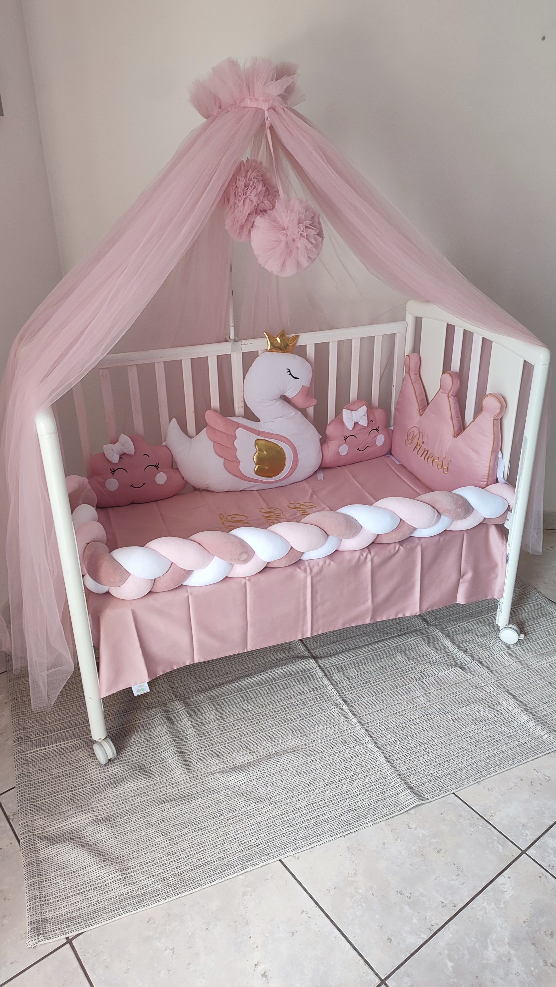 Set lettino neonata rosa con baldacchino in tulle, paracolpi a treccia e cuscini decorativi a forma di cigno e corona.