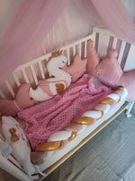 Set lettino neonata rosa con coperta lavorata a maglia, paracolpi a treccia bianco e oro e cuscini decorativi a cigno.