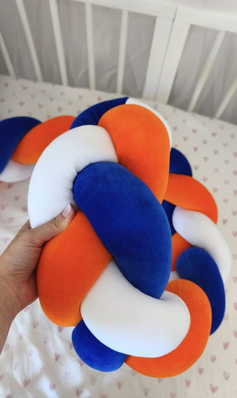 Mano che tiene la treccia paracolpi a 3 fili blu royal, arancione e bianco in velluto, dettaglio dell'intreccio morbido nel lettino