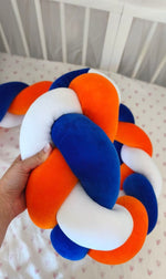 Mano che tiene la treccia paracolpi a 3 fili blu royal, arancione e bianco in velluto, dettaglio dell'intreccio morbido nel lettino
