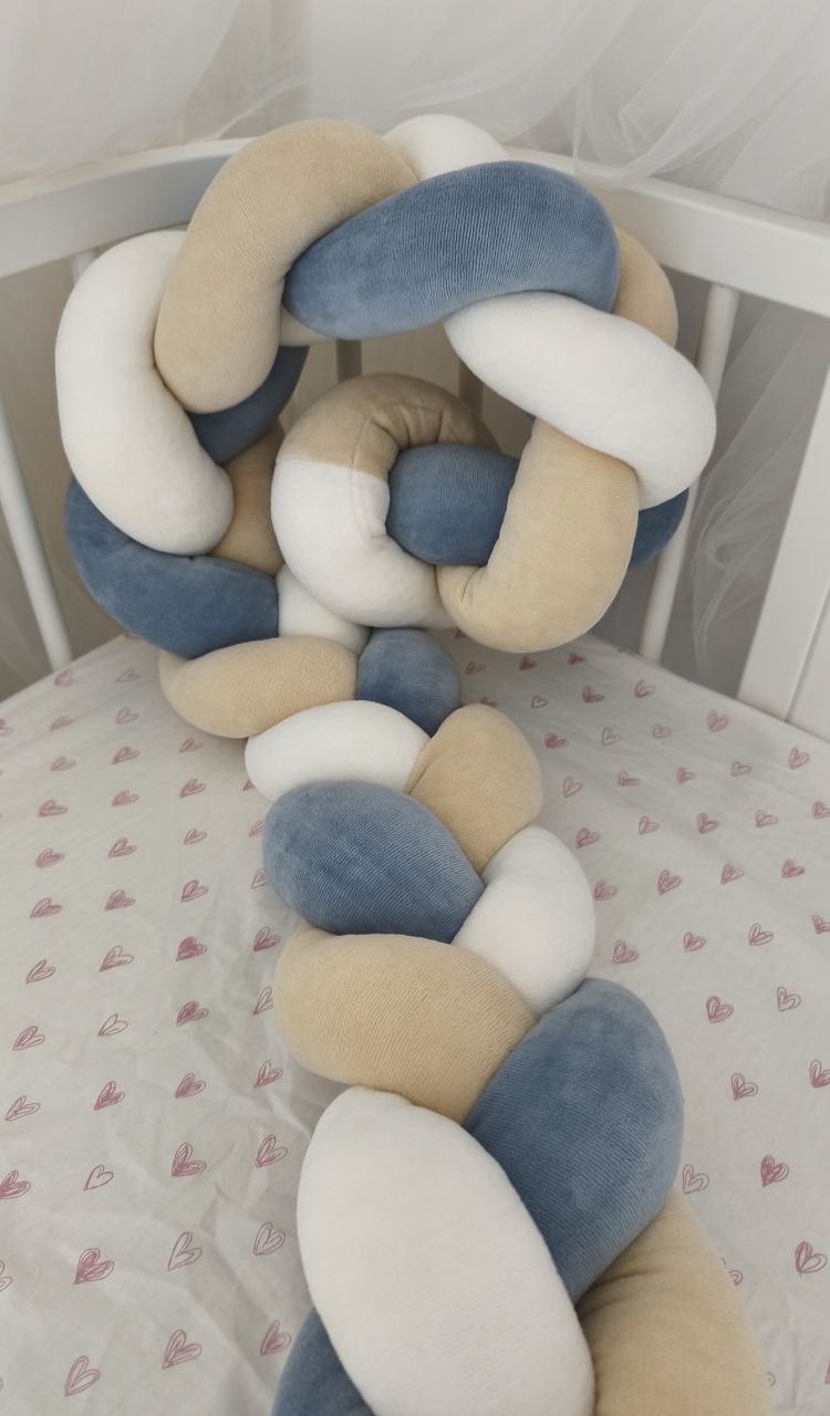 Treccia paracolpi a 3 fili blu polvere, beige classico e bianco in velluto, avvolta dentro un lettino ovale bianco con lenzuolino a cuori