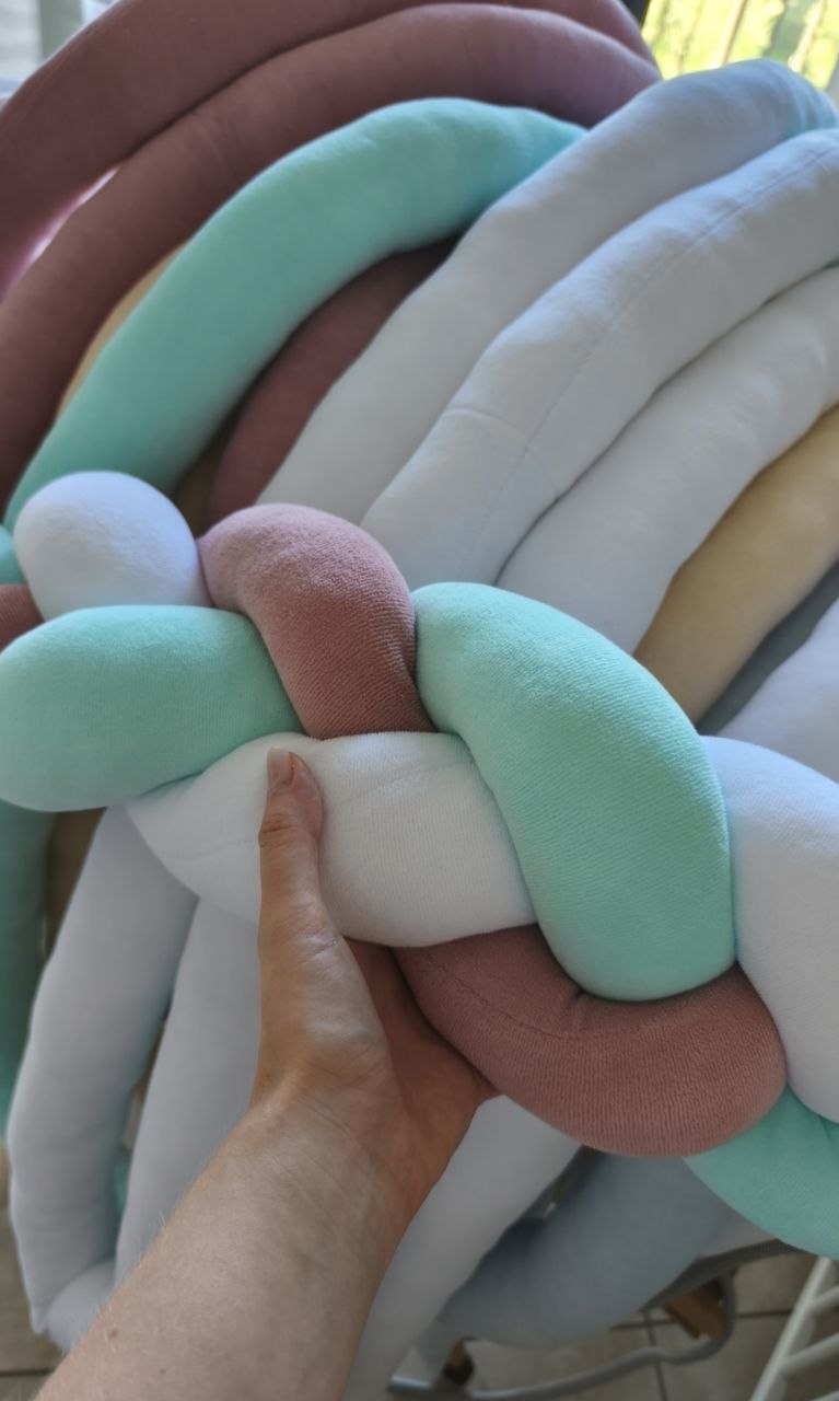 Mano su trecce paracolpi in velluto menta, rosa antico e bianco, dettaglio della morbidezza dell'imbottitura
