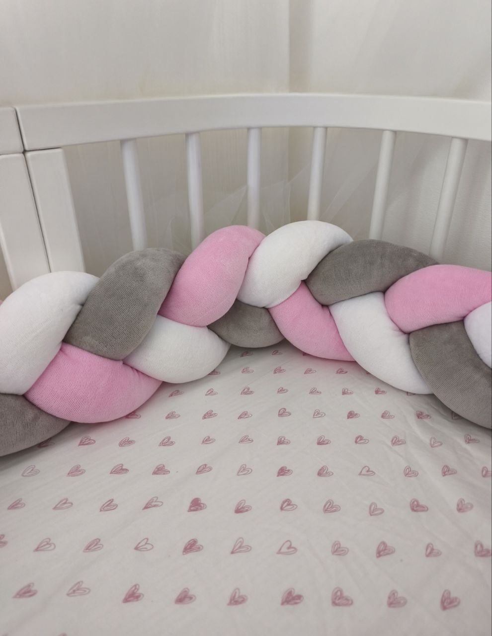 Treccia paracolpi rosa baby, grigio classico e bianco montata intorno al lettino con lenzuolo a cuoricini