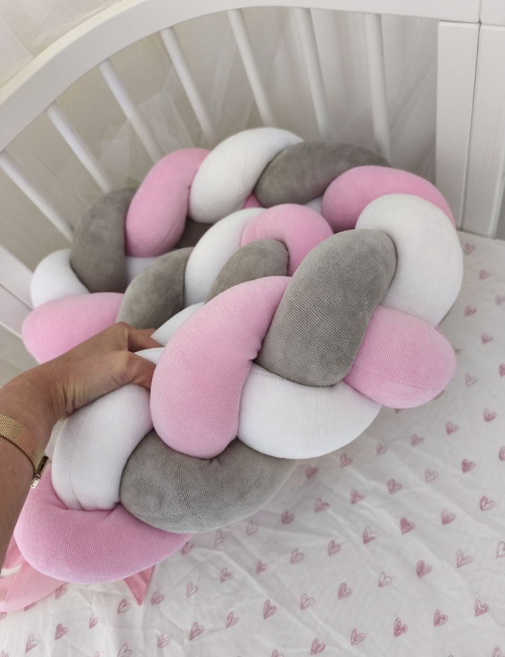 Mano che tiene la treccia paracolpi a 3 fili rosa baby, grigio classico e bianco in velluto, dettaglio dell'intreccio morbido nel lettino